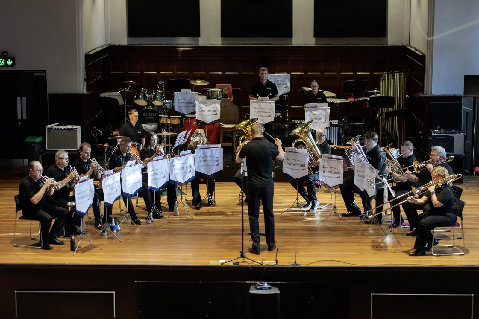 A large musical ensemble is performing on stage with a conductor in the center, featuring musicians playing saxophones, trombones, and percussion instruments. The stage has wooden flooring and a dark wood paneled backdrop, with music stands displayin