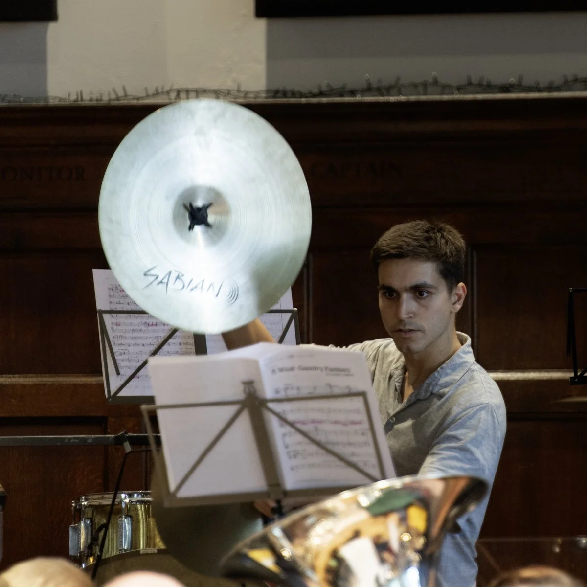 A young man with short brown hair playing a cymbal in an indoor music setting, with music stands and sheet music in front of him.