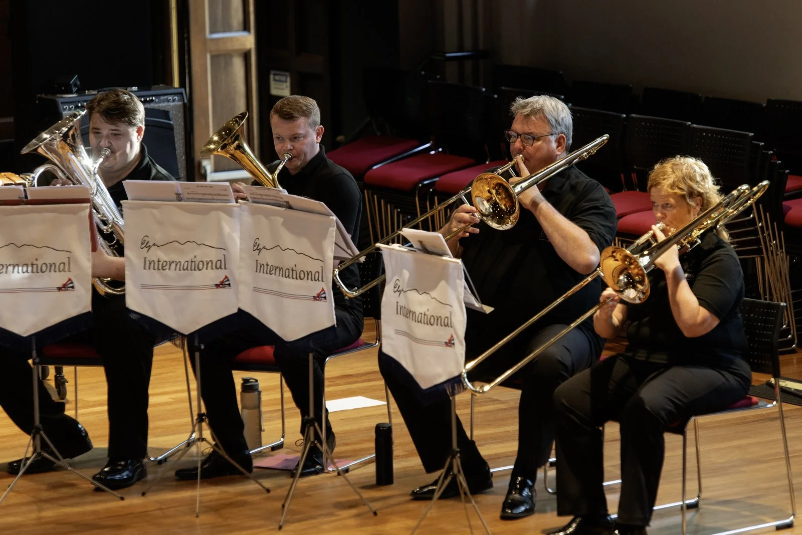 A group of four musicians sitting in a concert hall, playing brass instruments such as trombones and euphoniums, with music stands labeled 'International' in front of them.