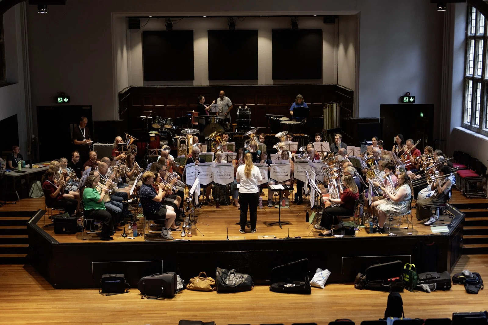 Orchestra rehearsing in a concert hall with musicians playing brass, woodwind, and percussion instruments, under the direction of a conductor.