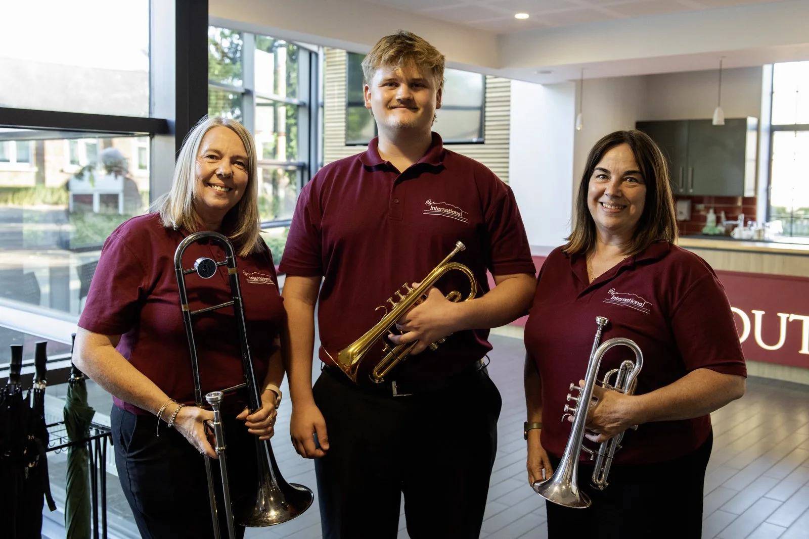 Three women and one young man standing indoors, all wearing matching maroon shirts, holding musical instruments including a trombone, trumpet, and possibly a saxophone, smiling at the camera.
