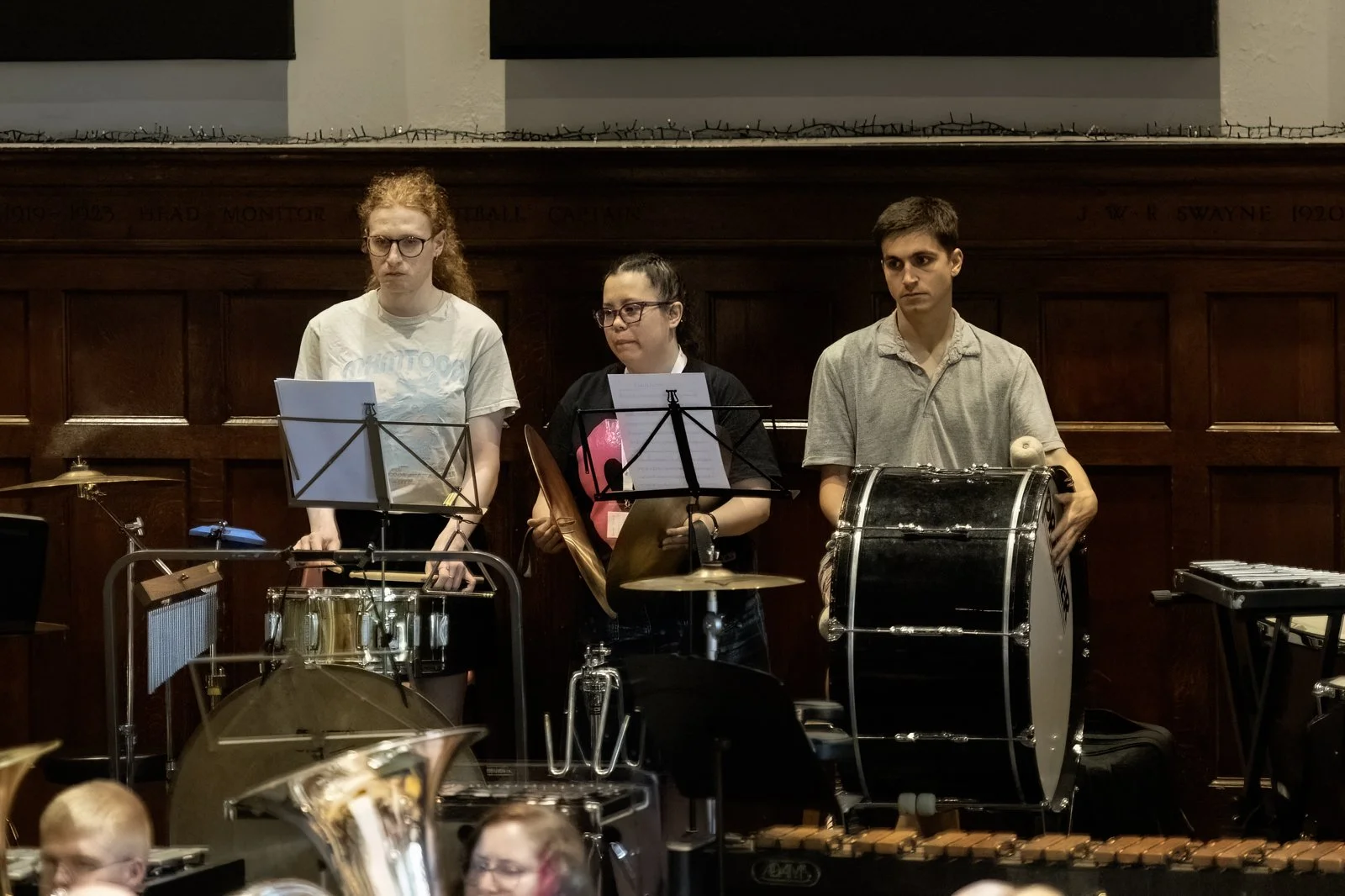 Three young musicians standing behind their instruments, which include a snare drum, a bass drum, and a xylophone, in a performance hall with wooden paneling.