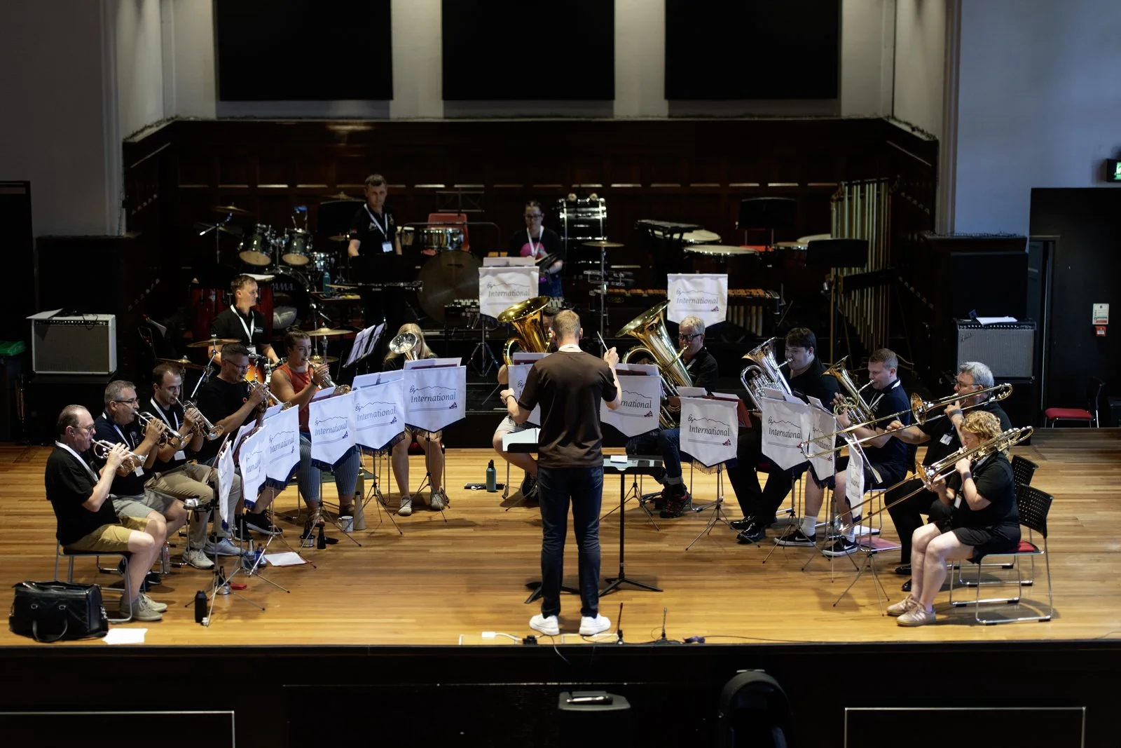 An orchestra performing on stage, conducted by a conductor standing with back to the camera. The orchestra includes brass, woodwind, and percussion instruments, with music stands labeled 'International.' The stage has a wooden floor and a dark backdr