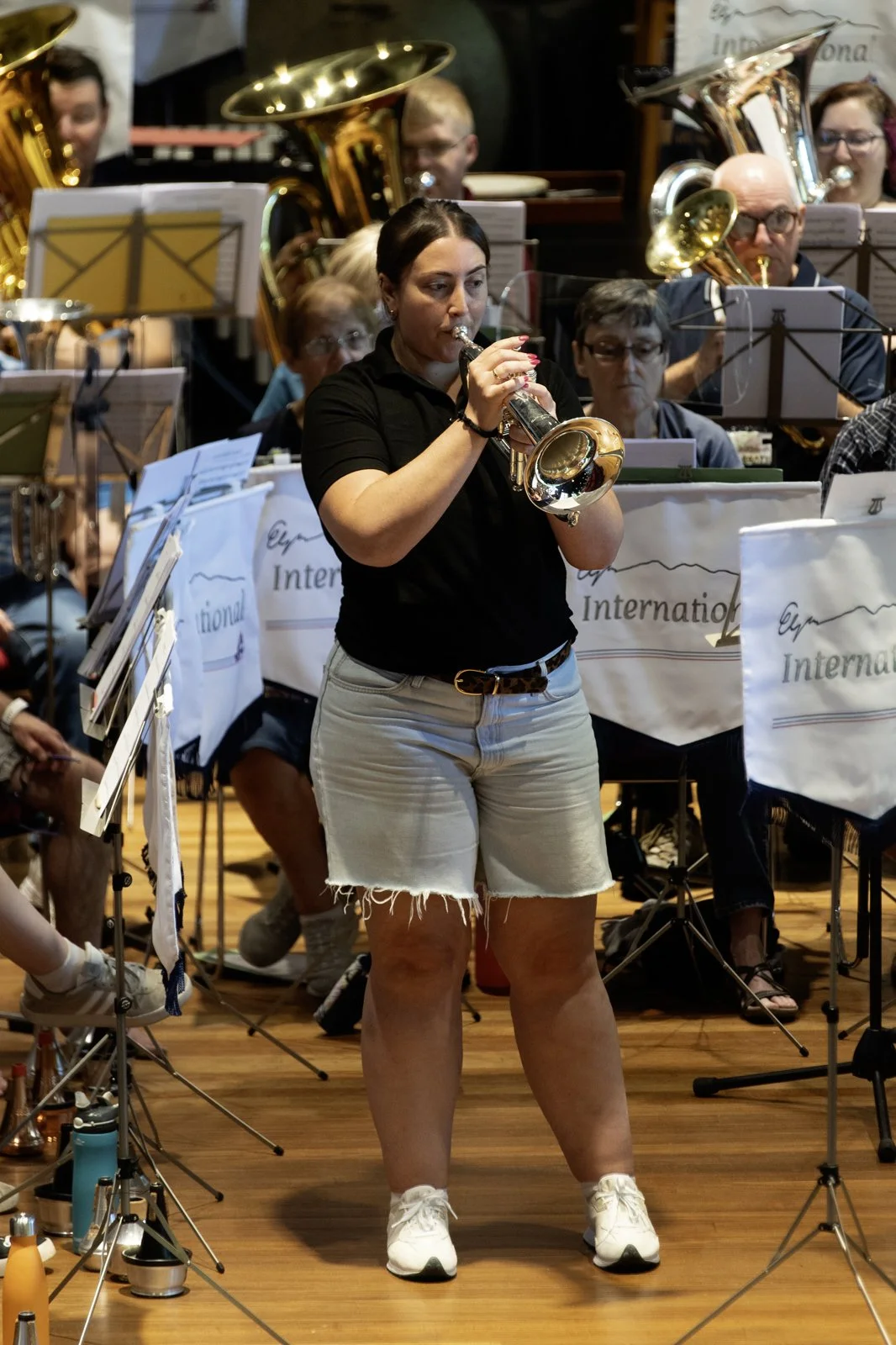 A woman in a black shirt and white shorts playing a trumpet during a band rehearsal, with other musicians and sheet music stands in the background.