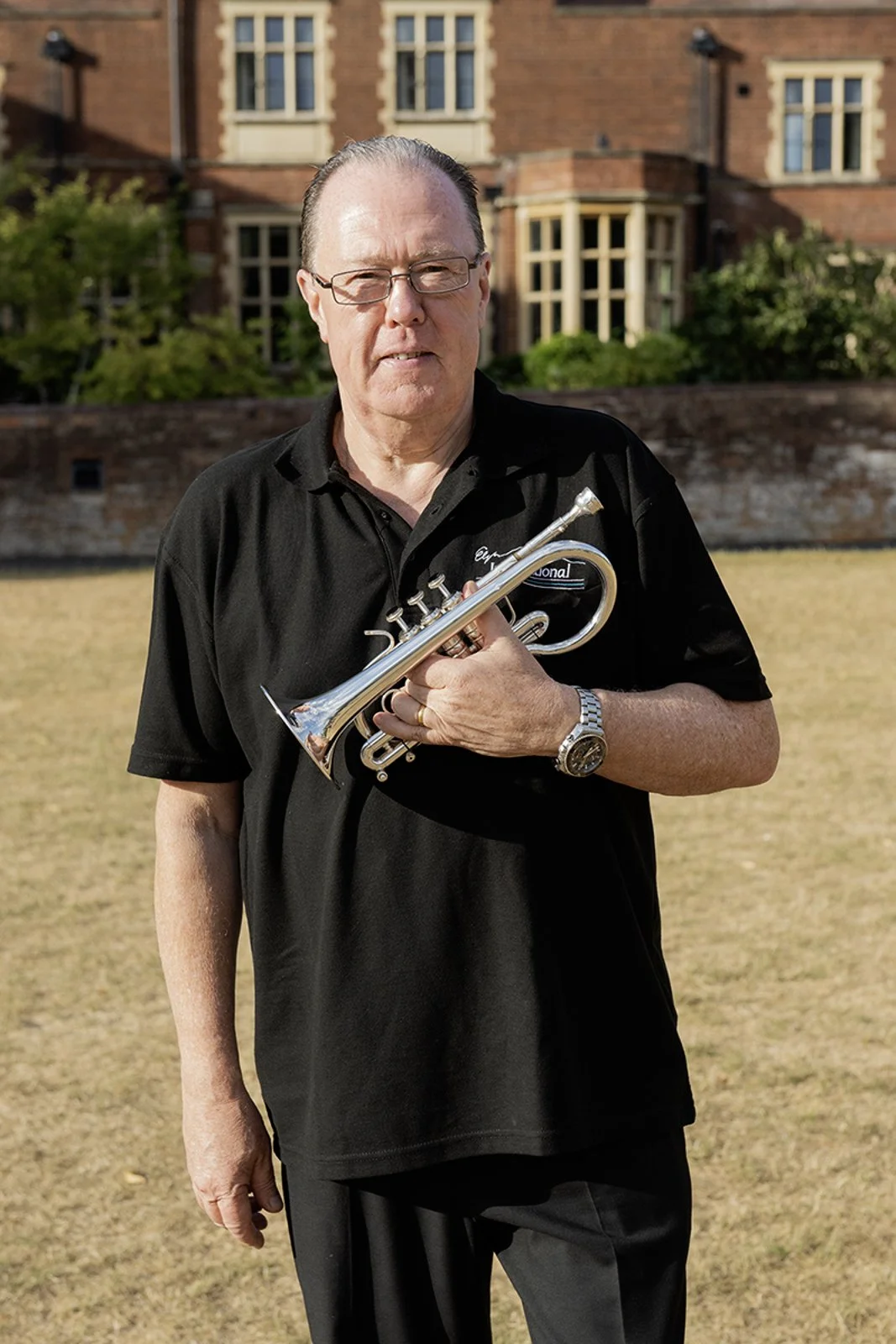 Older man with glasses holding a silver trumpet, standing on a grassy area with large brick building in the background.