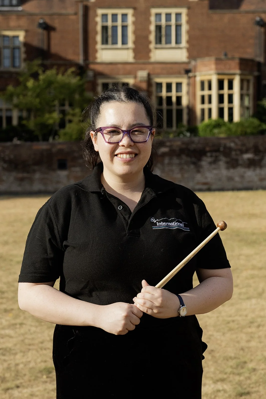 A woman with glasses and dark hair holding a drumstick outdoors in front of a historic brick building.