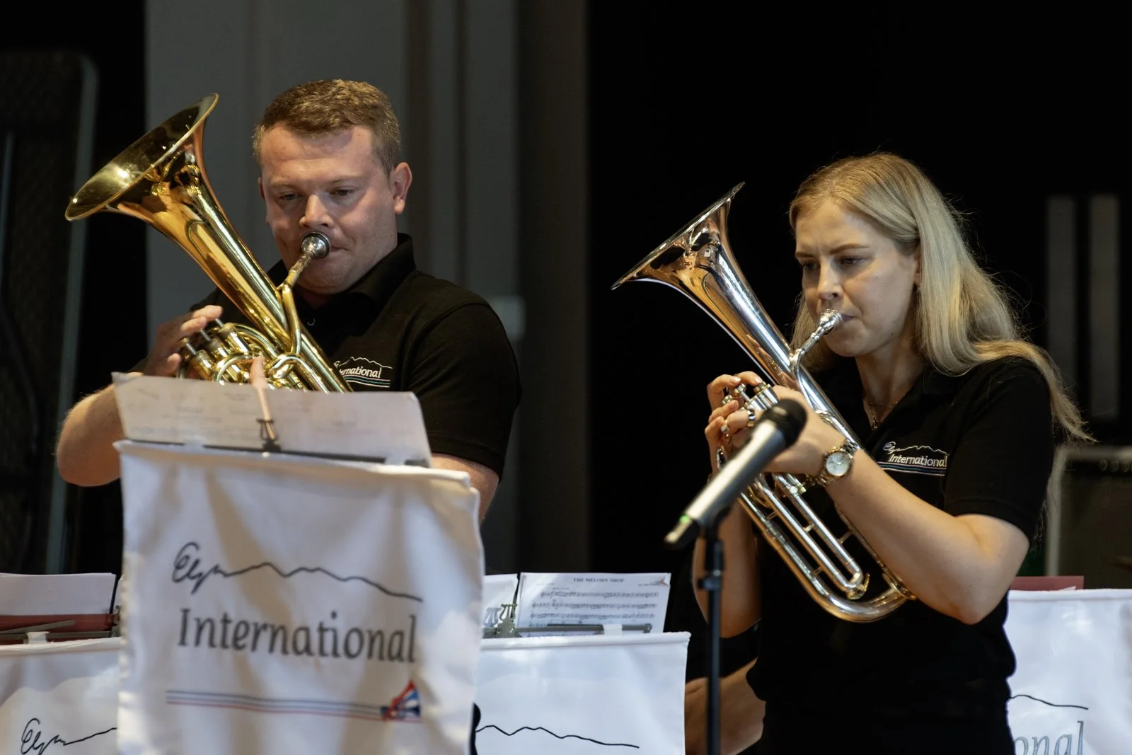 Two musicians playing trumpets during a performance, with sheet music in front of them and a microphone nearby.