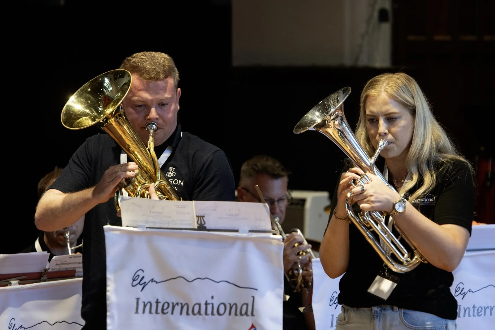Two musicians, a man and a woman, playing brass instruments during a concert. They are reading sheet music, with other musicians visible in the background.