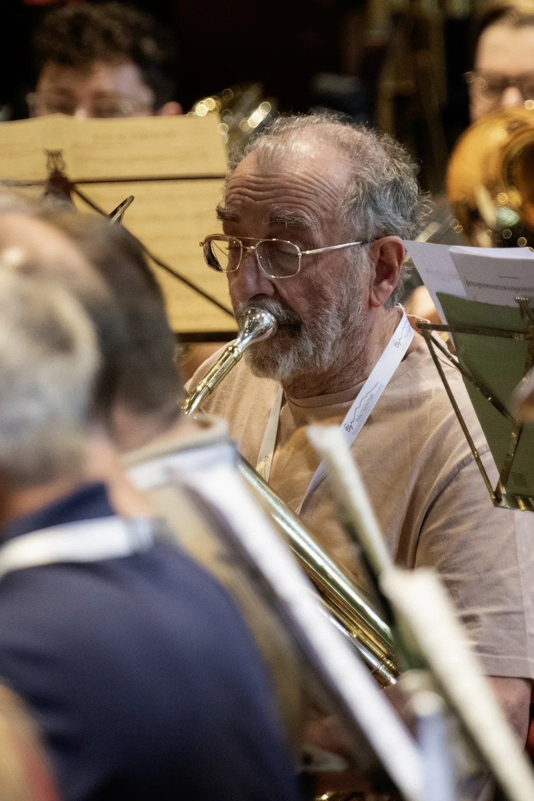 An older man with glasses and a beard playing a brass instrument in an orchestra, with other musicians around him, in a concert setting.