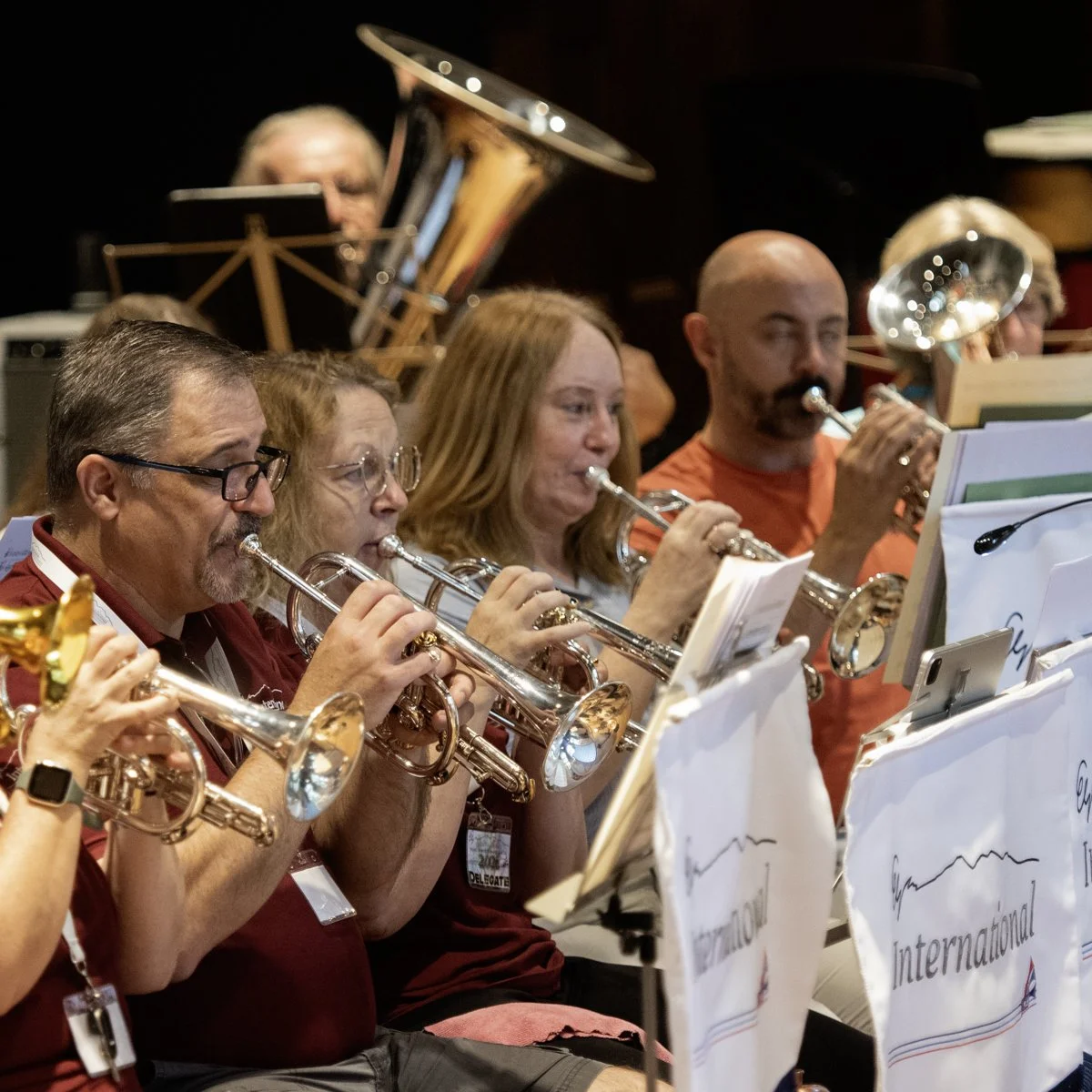 A group of different age people playing brass instruments during an orchestra rehearsal or concert, with sheet music in front of them.