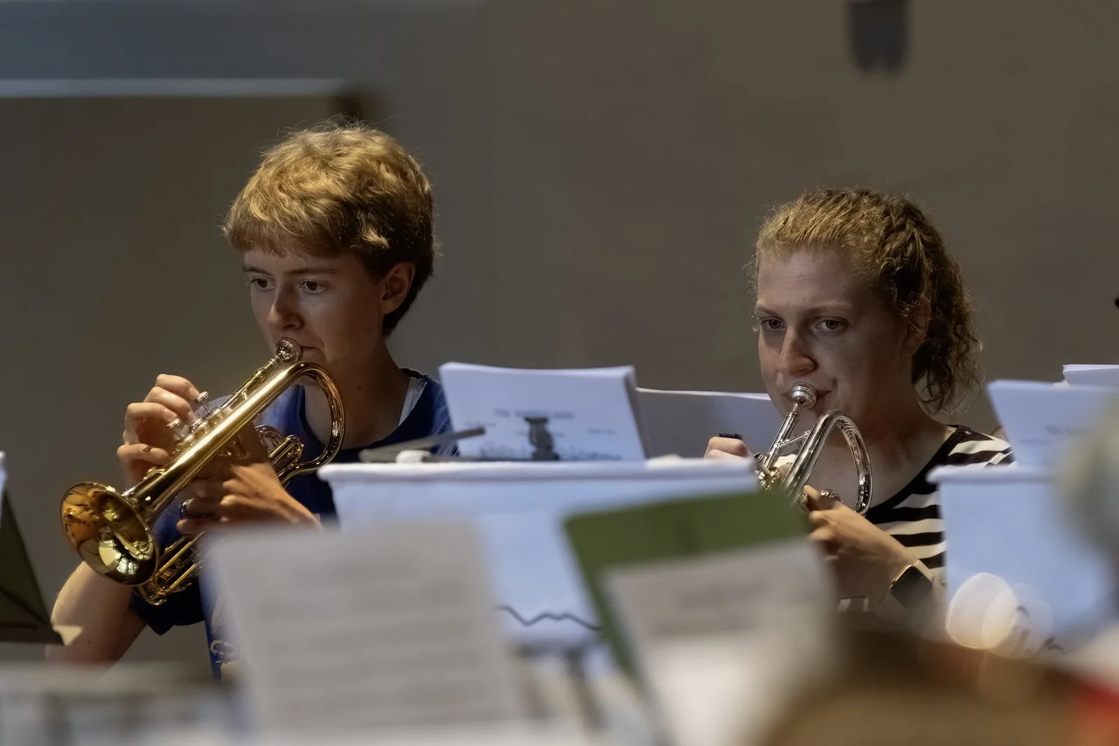 Two women playing trumpets during a music rehearsal, surrounded by sheet music and music stands.