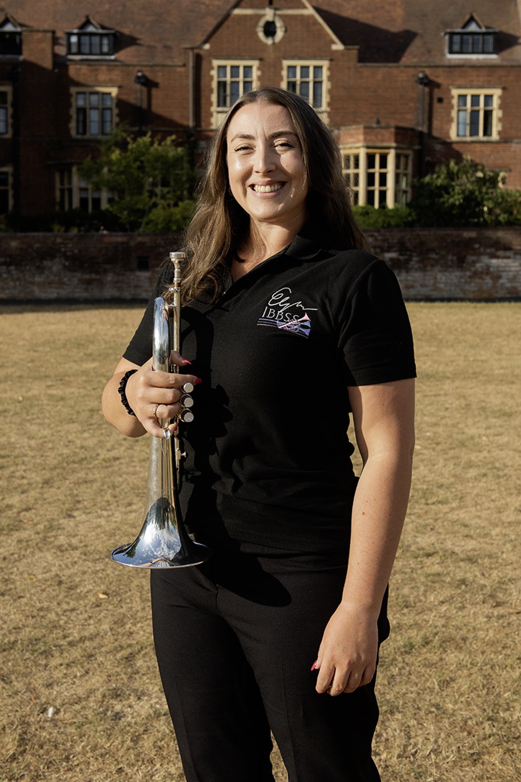 A woman smiling outdoors holding a trumpet in one hand. She is wearing a black polo shirt with a logo and text on it. A large brick house with many windows is in the background.