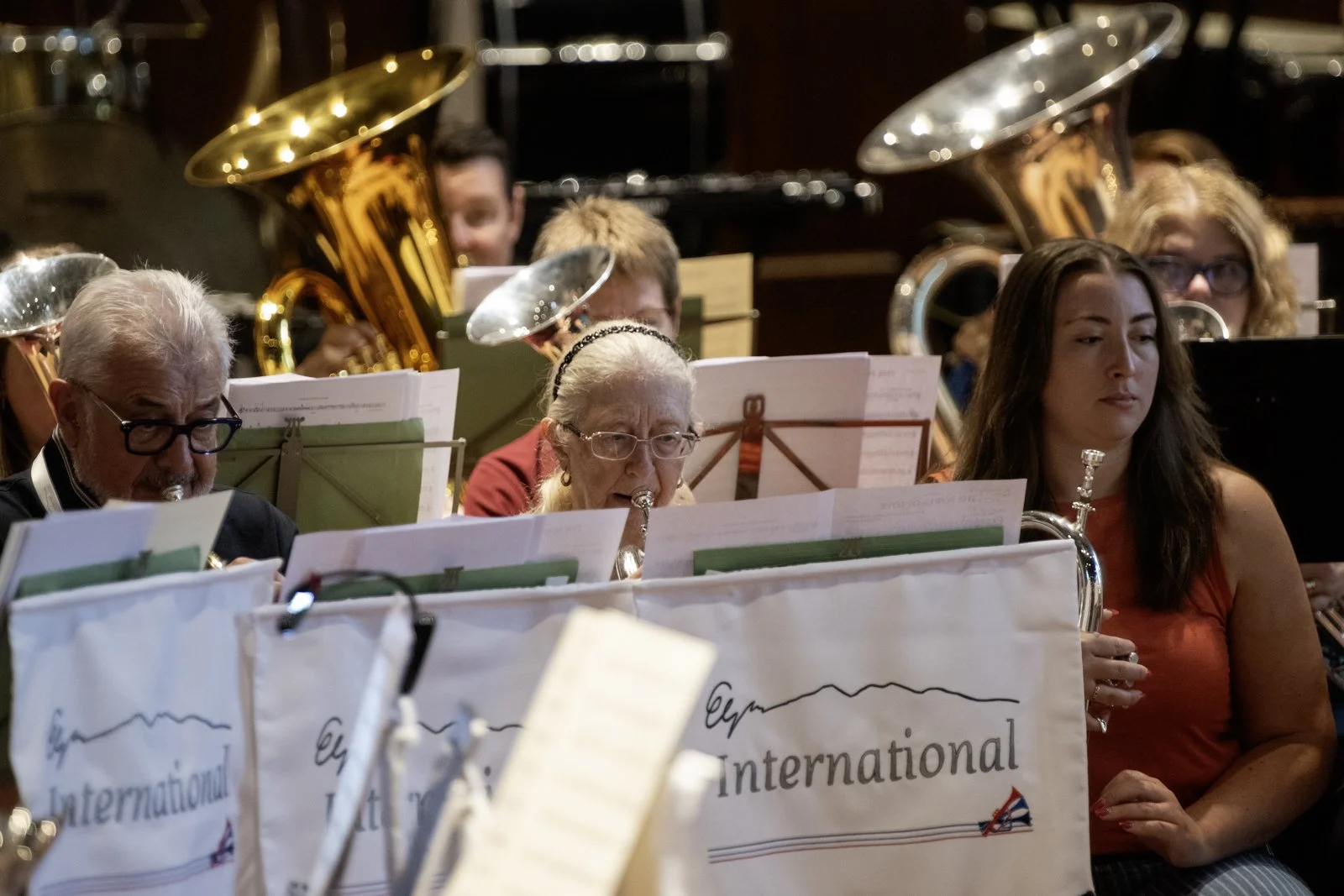 An orchestra performing, with musicians playing brass instruments; music stands with sheet music in foreground; some musicians appear to be looking at their music, others are focused on playing.