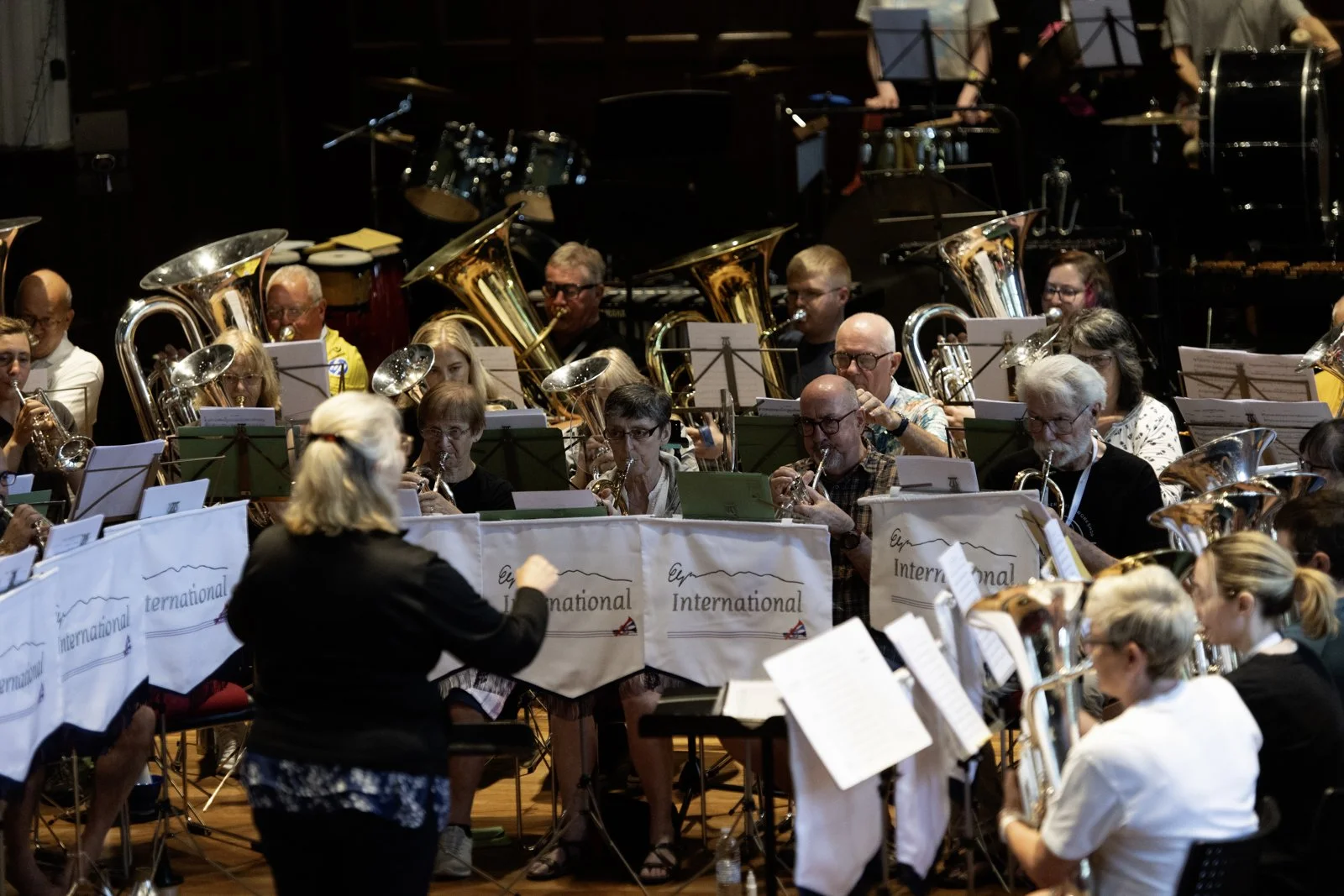 An orchestra performing on stage, with conductors, musicians, and a variety of brass and wind instruments, during a concert.