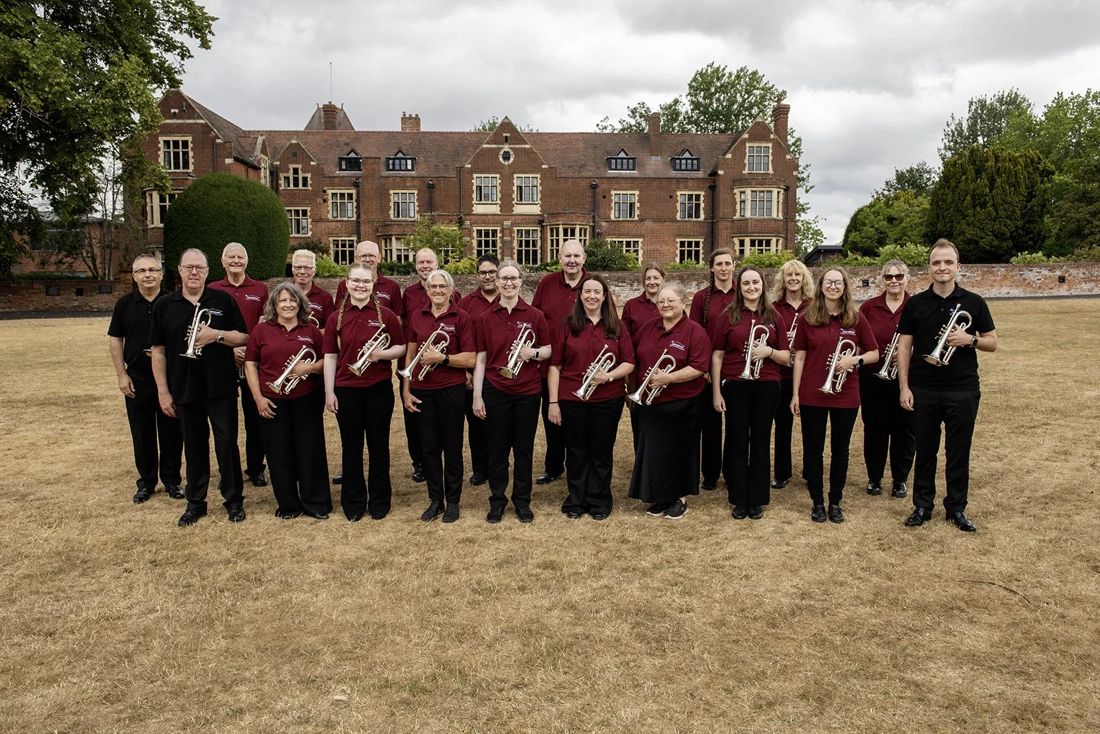 Group of people in matching maroon shirts, some holding trumpets, standing on a grassy field with a castle-like building in the background.