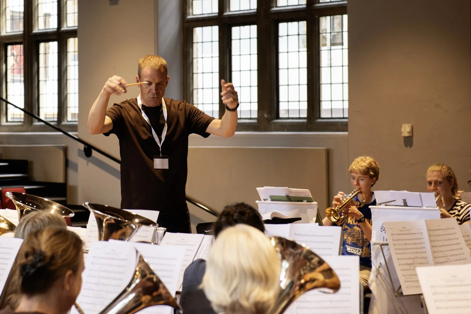 An orchestra rehearsal with a conductor leading young musicians in a room with large windows.