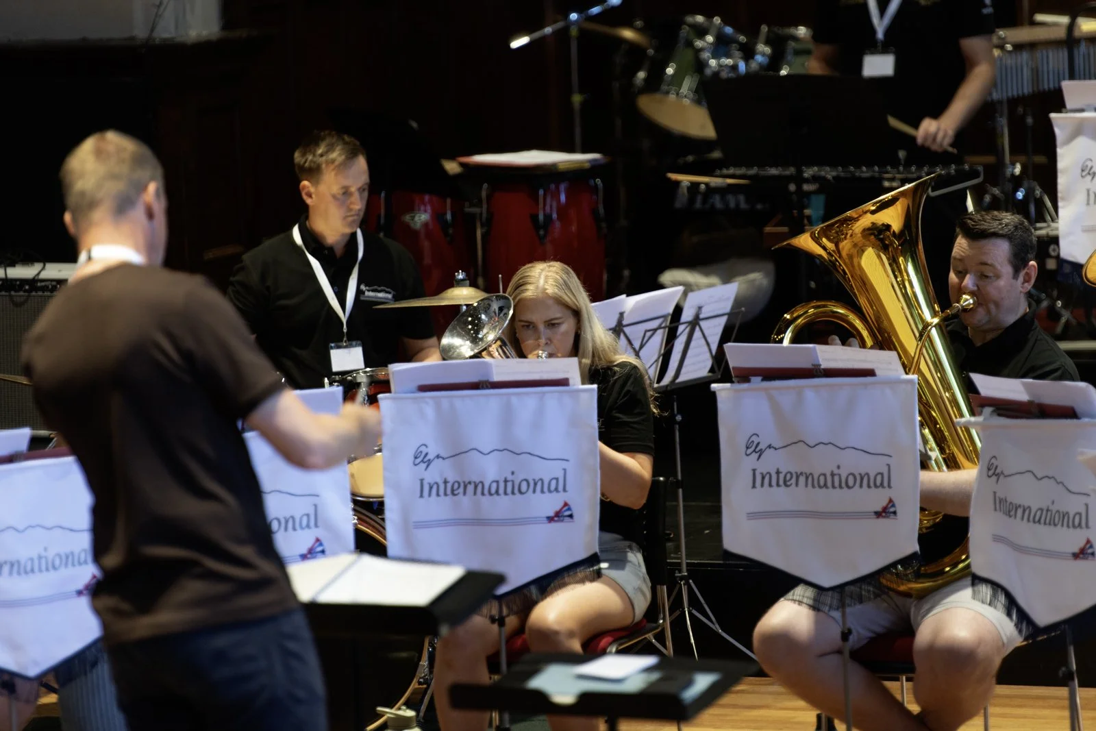 A group of musicians performing in a band, with some playing wind instruments and others, including a person in the background, standing behind percussion instruments, during a concert or rehearsal.