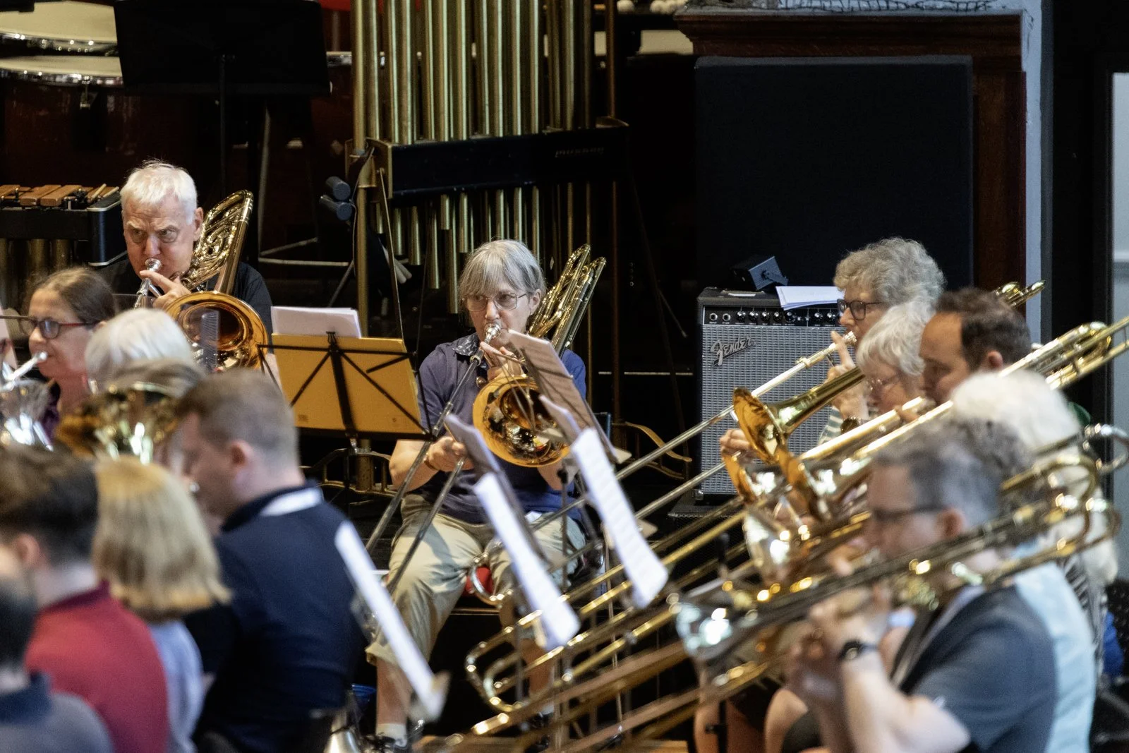 An orchestra performing indoors with musicians playing wind instruments, including saxophones, trumpets, and clarinets, with sheet music stands and a Fender amplifier visible in the background.