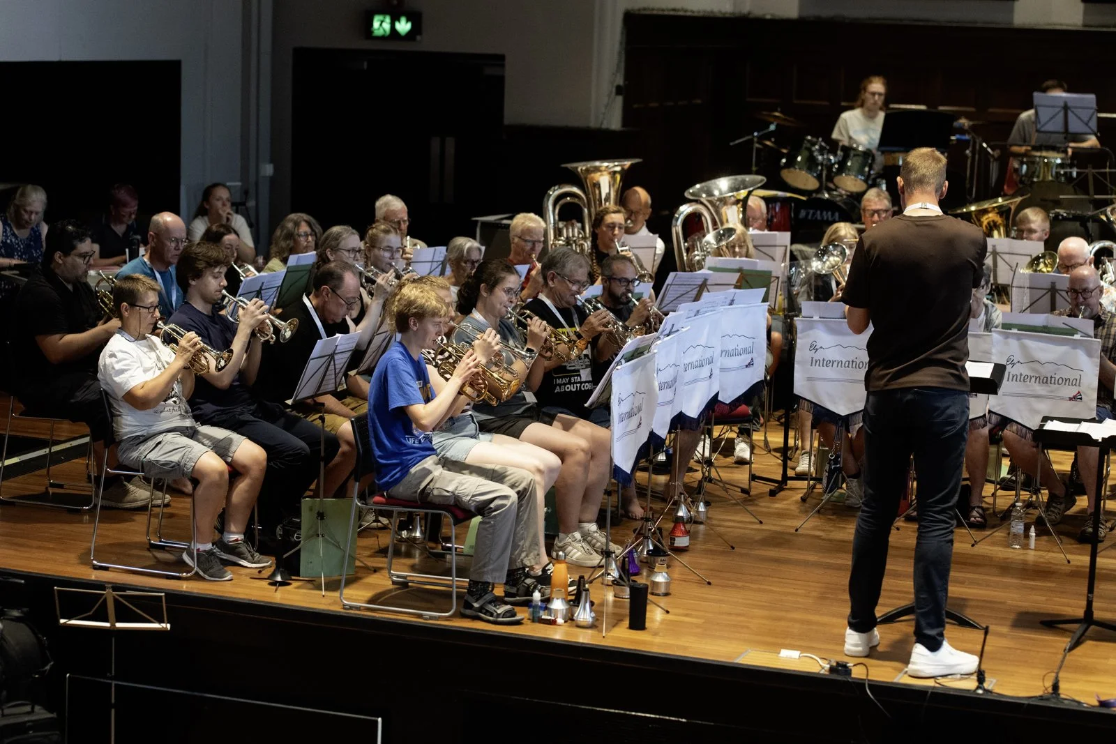 An orchestra performing on stage with a conductor standing in front of them. The musicians are playing various wind instruments. The stage has wooden flooring and music stands with banners that read 'International.' In the background, there are music