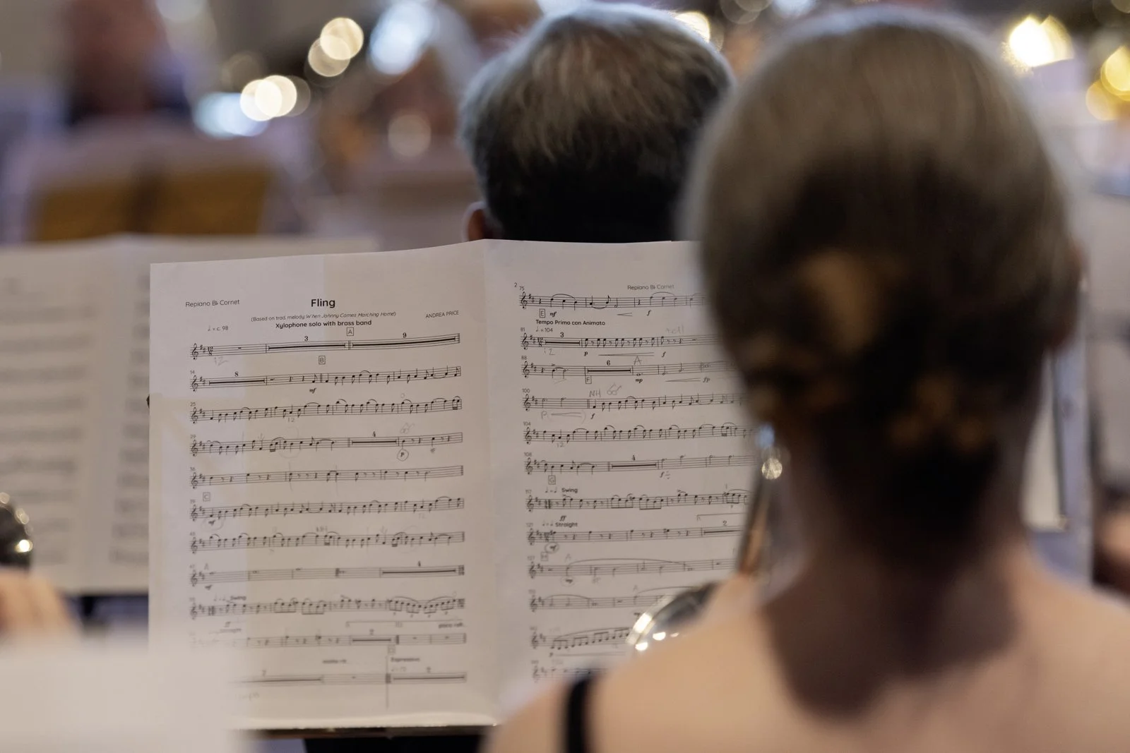 A woman and a man are seated in an orchestra, reading sheet music for a brass band piece titled "Fling" by Andrea Price. The background is blurred with warm and out-of-focus lights.