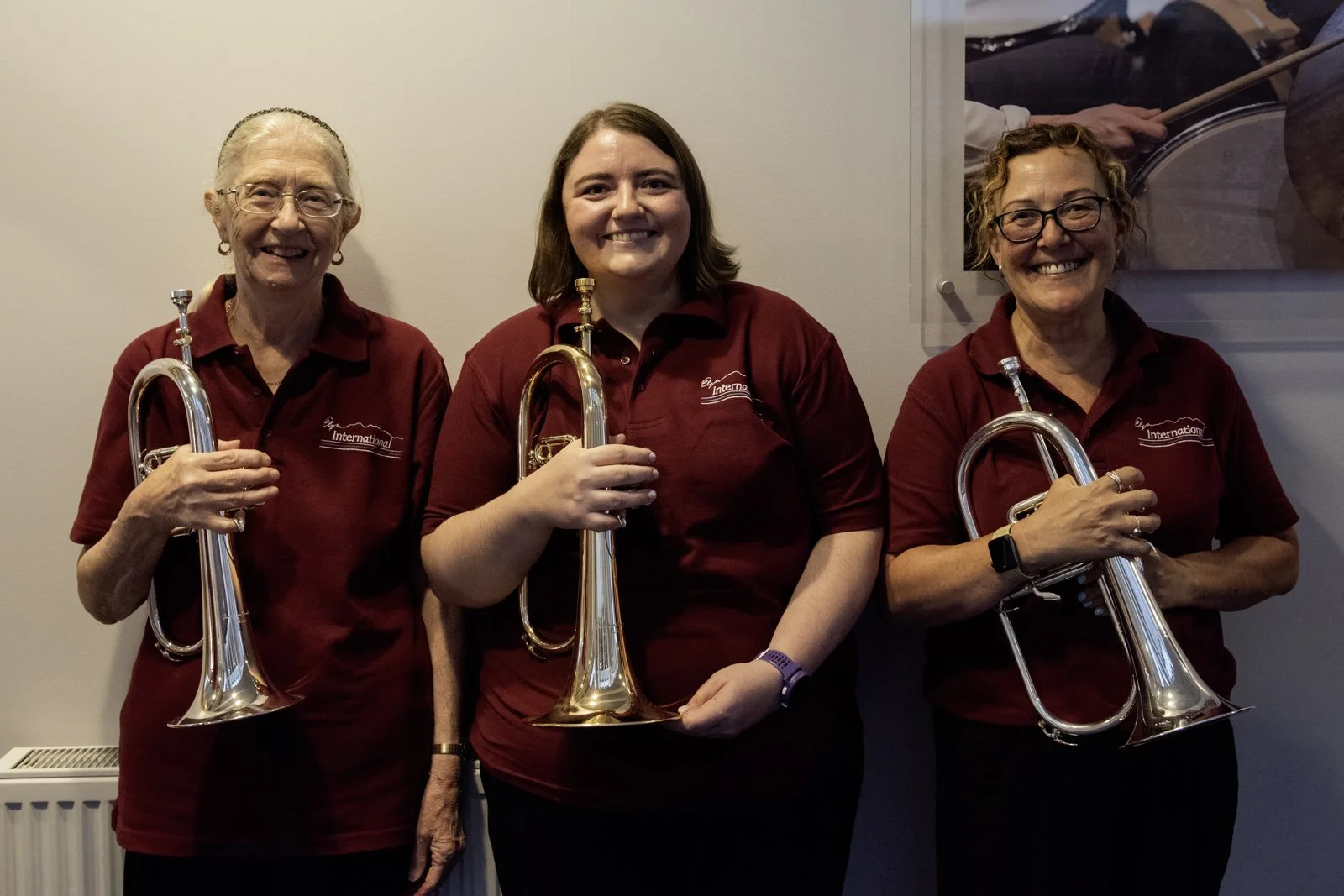 Three women standing side by side, each holding a brass musical instrument, possibly a trumpet, inside a room with plain walls and a framed photo of a drummer in the background. All three women are smiling and wearing maroon polo shirts with a logo.