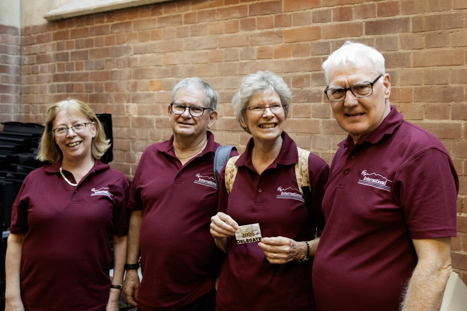 Four elderly people standing against a brick wall, wearing maroon shirts with a logo, smiling. One woman in the center holds a delegate badge.