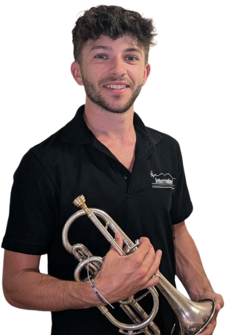 A young man with light brown hair, blue eyes, and facial hair, smiling and holding a trumpet, wearing a black polo shirt with a logo reading 'Intermountain' against a plain white background.