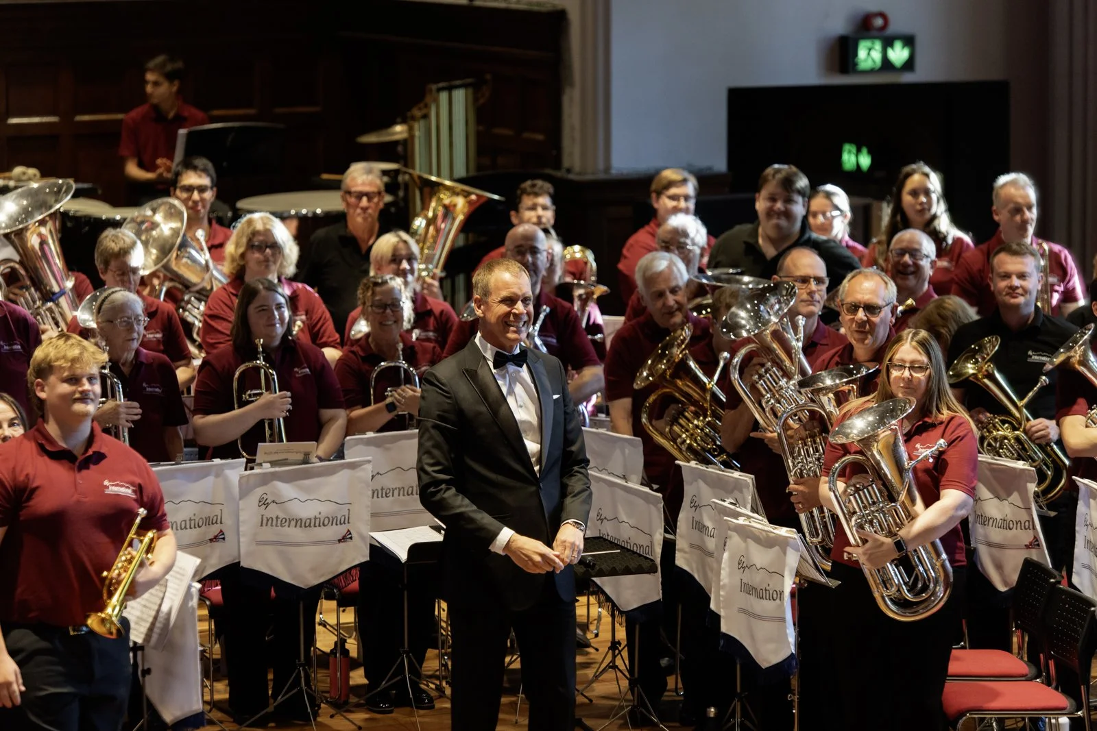 A conductor in a tuxedo stands at the front of a large orchestra with various musicians holding brass and woodwind instruments. The musicians are wearing matching maroon shirts, and the orchestra is organized behind white music stands with the words "International" printed on them. The group appears to be smiling and enjoying a performance or rehearsal.
