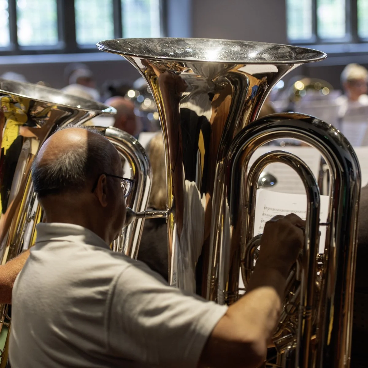 A man playing a brass instrument, likely a tuba, during a musical performance. There are other similar instruments and musicians visible in the background.