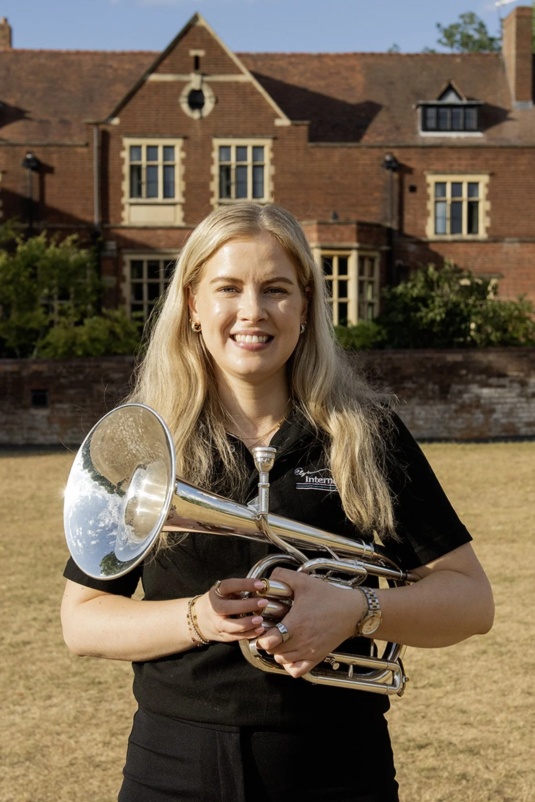A young woman with long blonde hair holding a silver trumpet, standing outdoors on a grassy area with a large brick building in the background. She is smiling and wearing a black shirt with a logo, jewelry, and a watch.