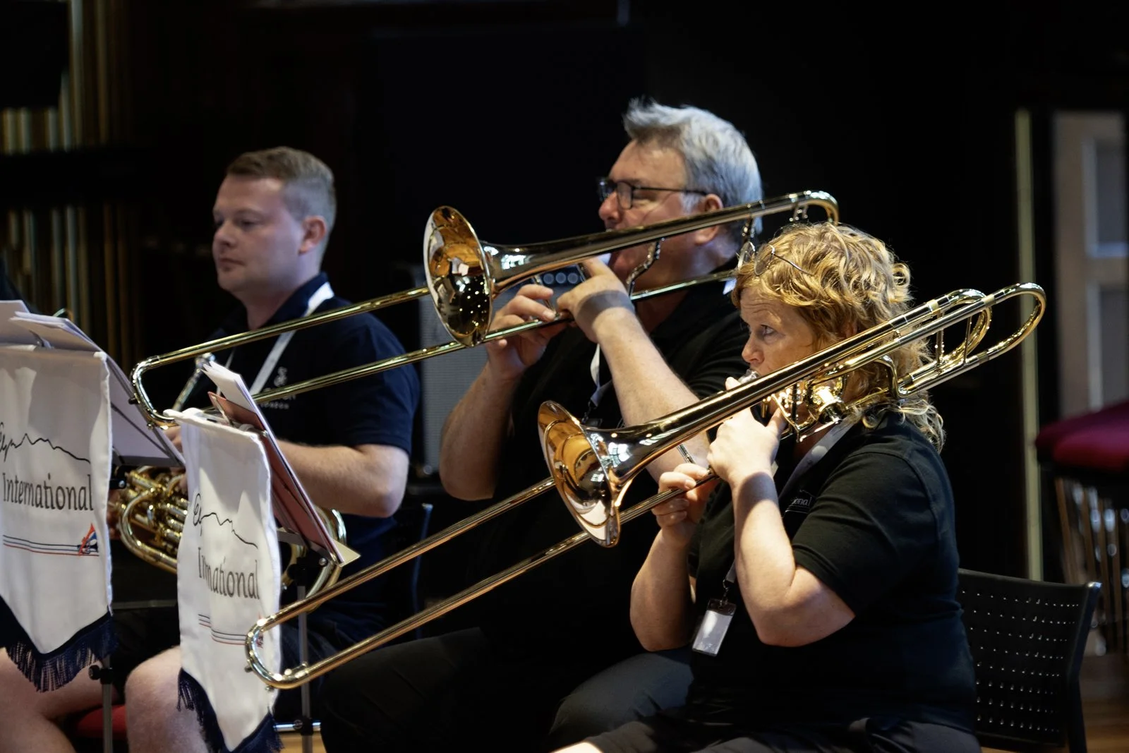 Three musicians playing trombones during a concert. One man and one woman are visible, reading sheet music, with music stands in front of them. The scene is indoors with a dark background.