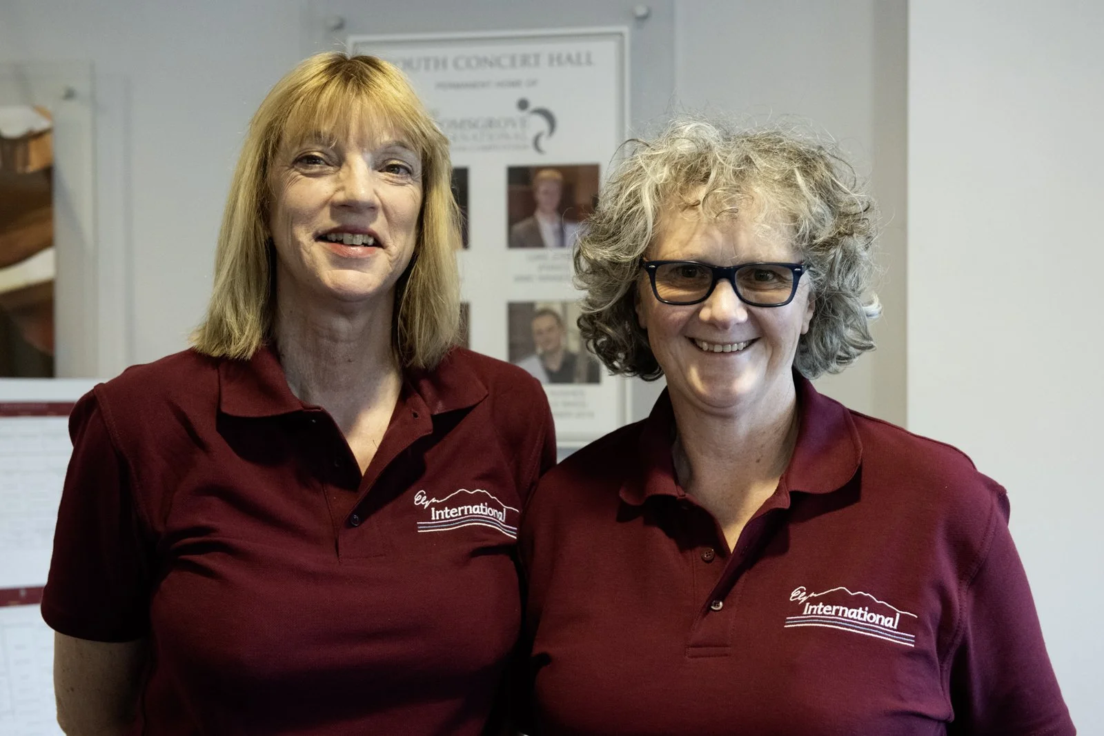 Two women wearing maroon shirts with 'International' logo stand indoors, smiling at the camera.