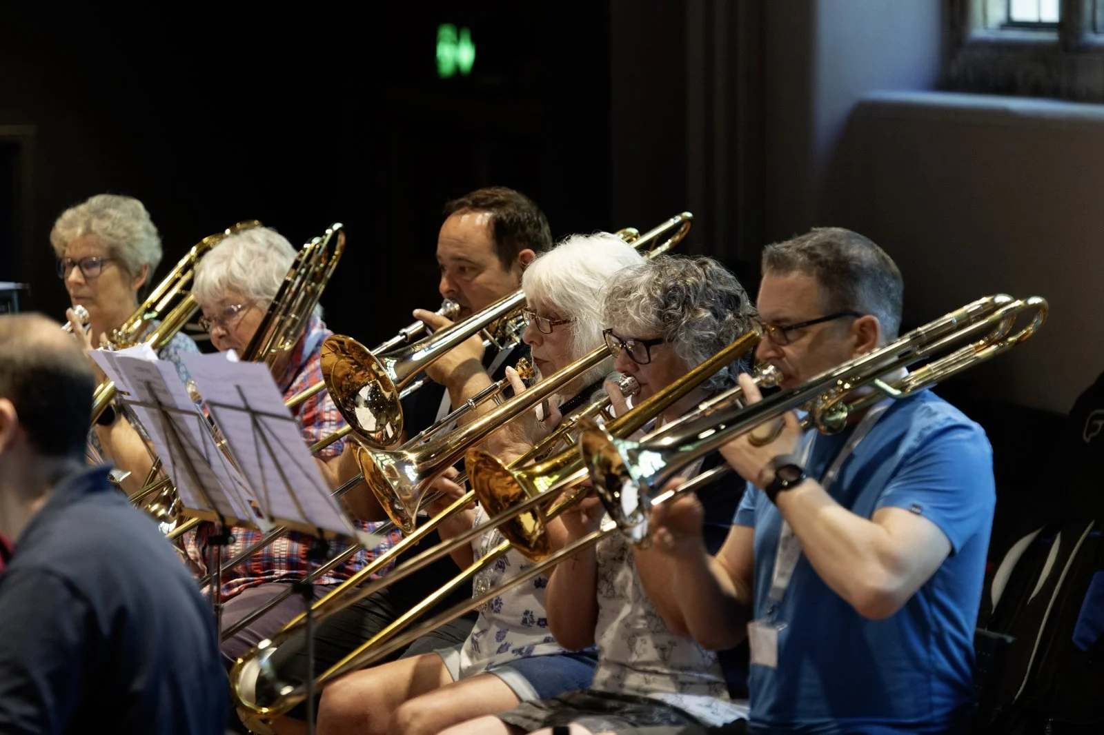Group of elderly and middle-aged musicians playing trumpets in an indoor setting, seated in a row, with music sheets in front of them.