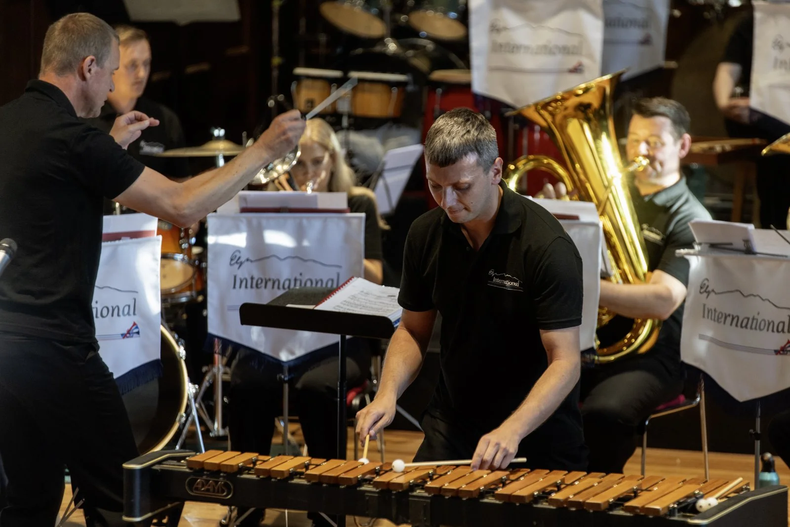 Musicians performing in a concert, featuring a xylophone and a brass instrument, with a conductor leading the ensemble.