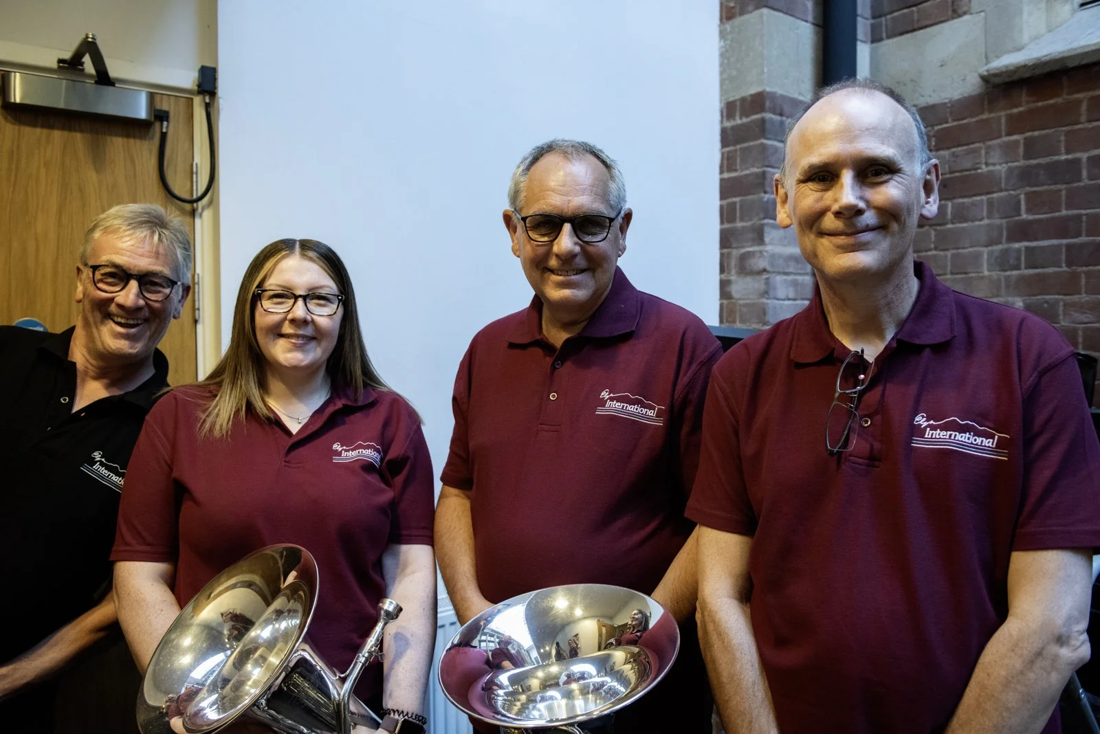 Four people standing together indoors, three men and one woman, smiling at the camera, with brass musical instruments in front of them, wearing maroon and black polo shirts with a logo.