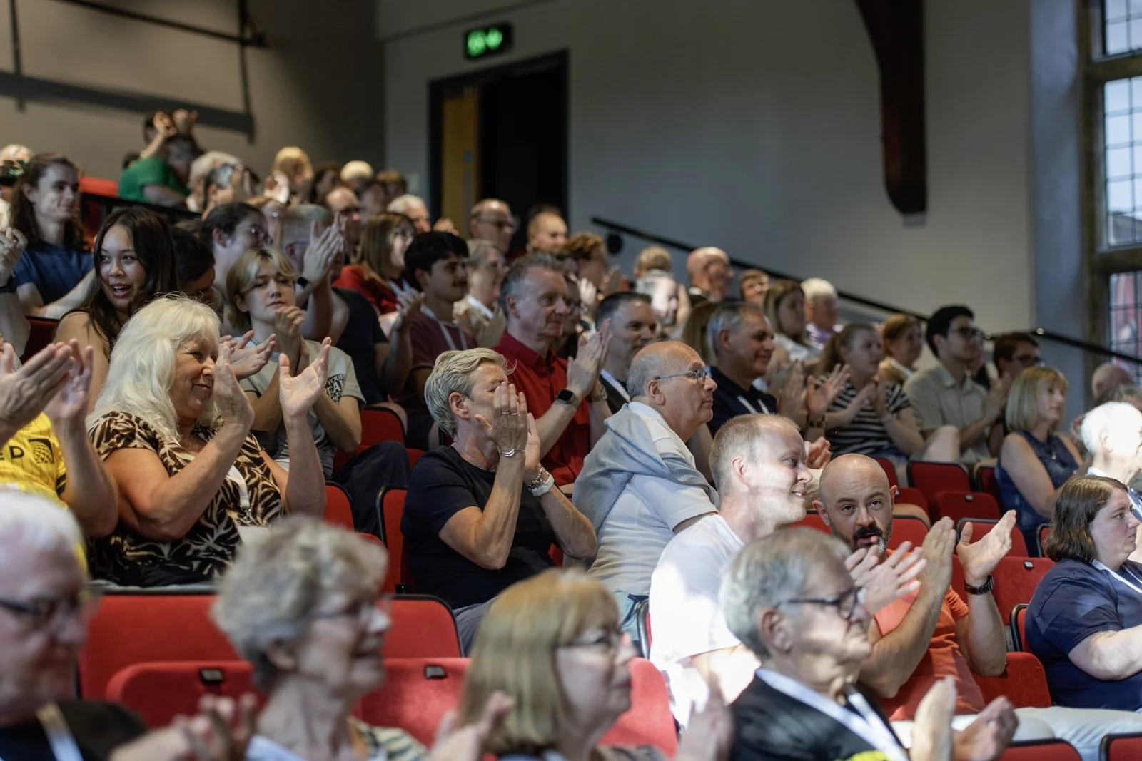 Audience members seated in red seats attending an event, clapping and smiling inside a large hall with high windows.
