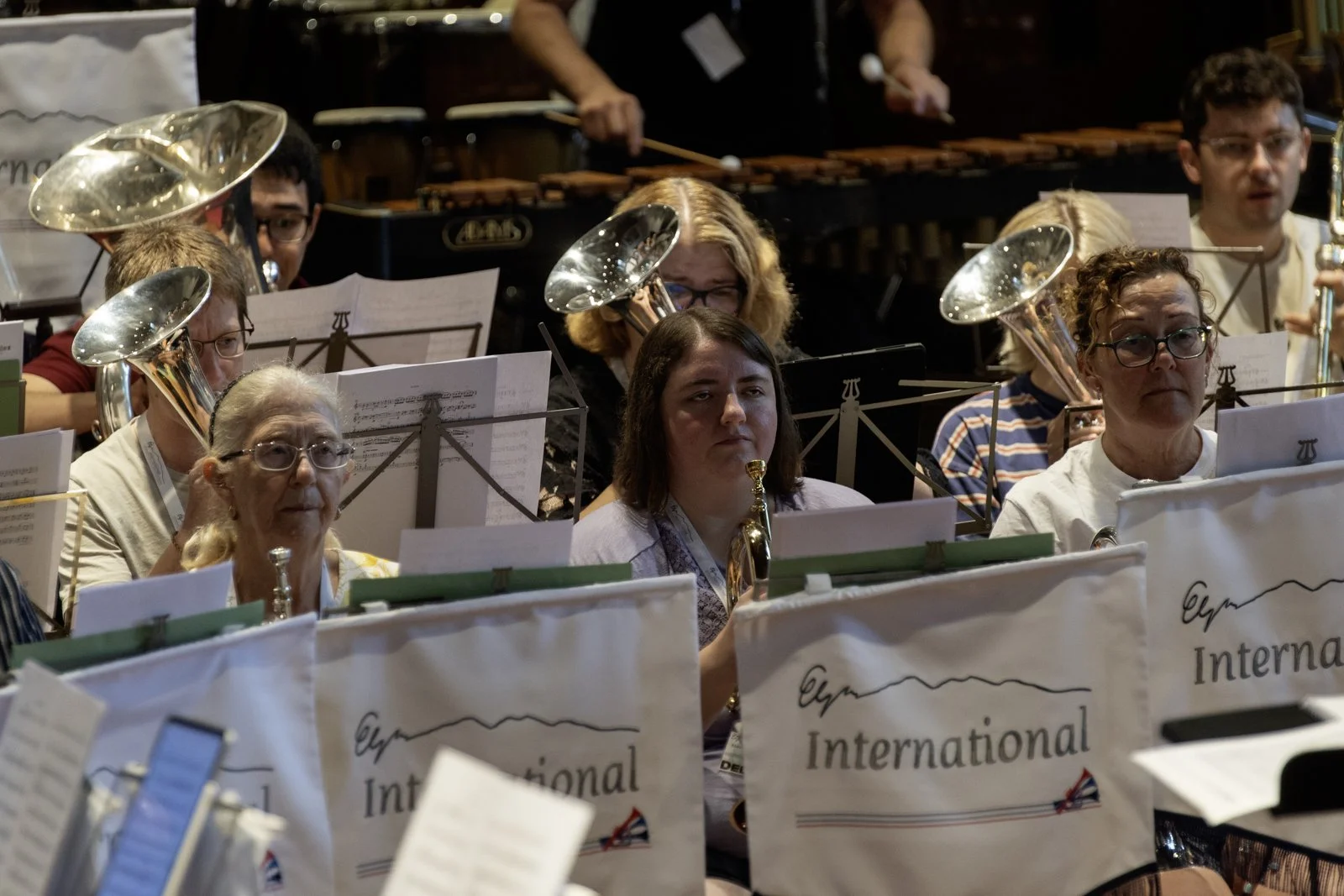 A group of musicians, mostly women, seated in a concert band setting with several brass and woodwind instruments, including tubas and saxophones, with music stands displaying the words 'International' at the top, performing in a dimly lit indoor venu