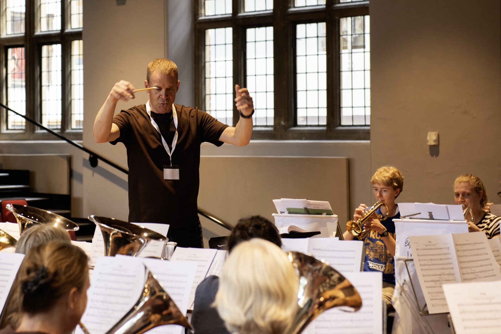 A music conductor leading an orchestra of children playing brass instruments, in a room with large windows.