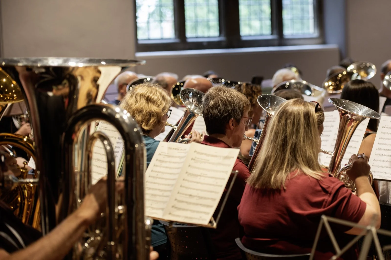 Orchestra of musicians playing wind instruments, with sheet music in front of them, in a room with large windows.
