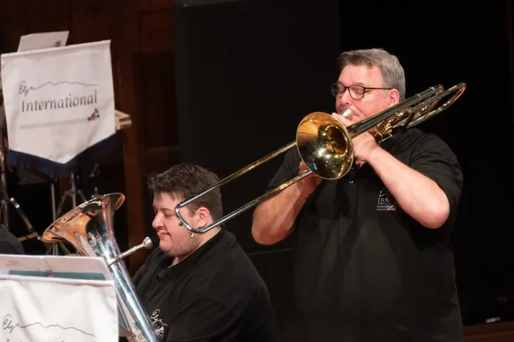 Two musicians playing brass instruments during a performance, one with a trombone and the other with a euphonium, in a concert hall.