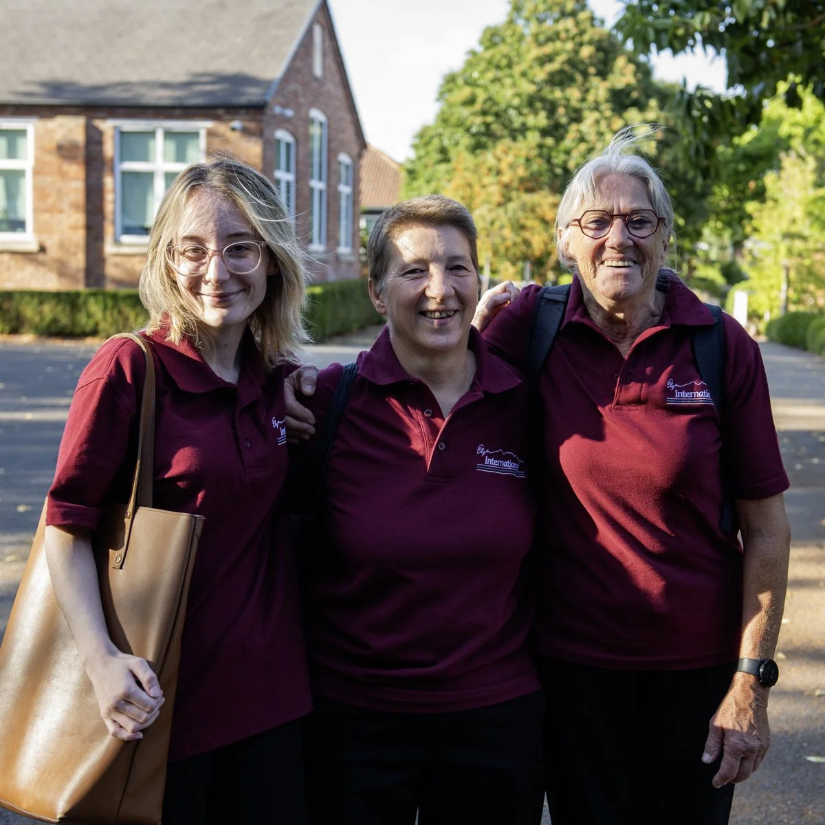 Three women standing outdoors, smiling, wearing matching maroon shirts with a logo on the left chest that says "International". They have their arms around each other, and the background features a brick building, green trees, and a sunny sky.