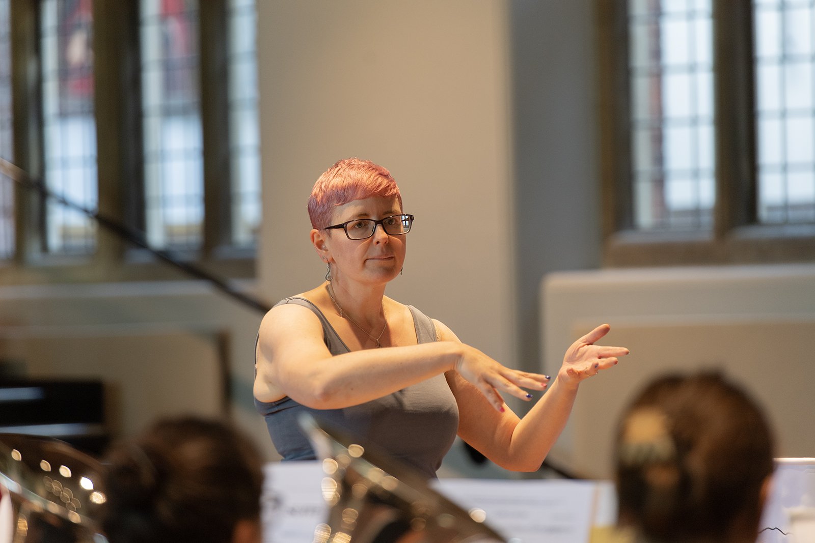 A woman with short pink hair and glasses conducting an orchestra in a room with tall windows.