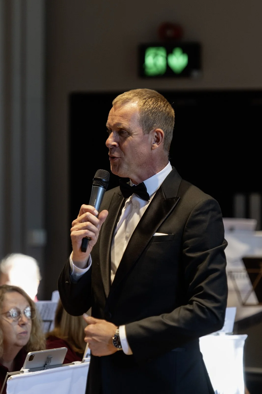 Man in a tuxedo speaking into a microphone at a formal event, with seated guests and a green emergency exit sign in the background.