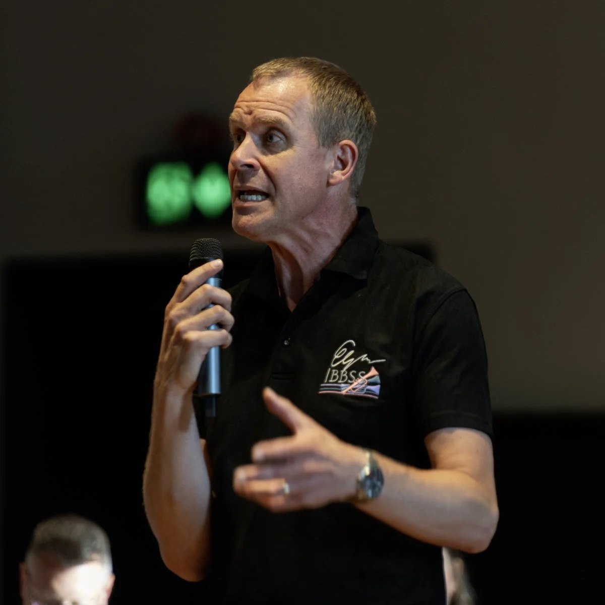 A man holding a microphone speaking at an event, wearing a black shirt with a logo that reads 'CYM IBSS'.