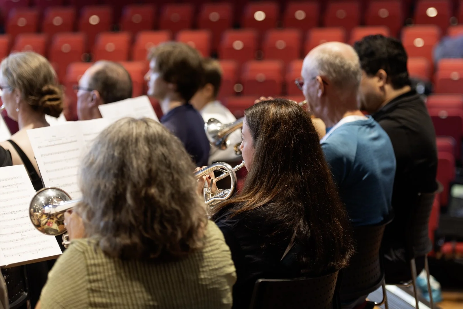A group of people participating in a musical ensemble, playing flutes and a trumpet, seated in a concert hall with red seats.