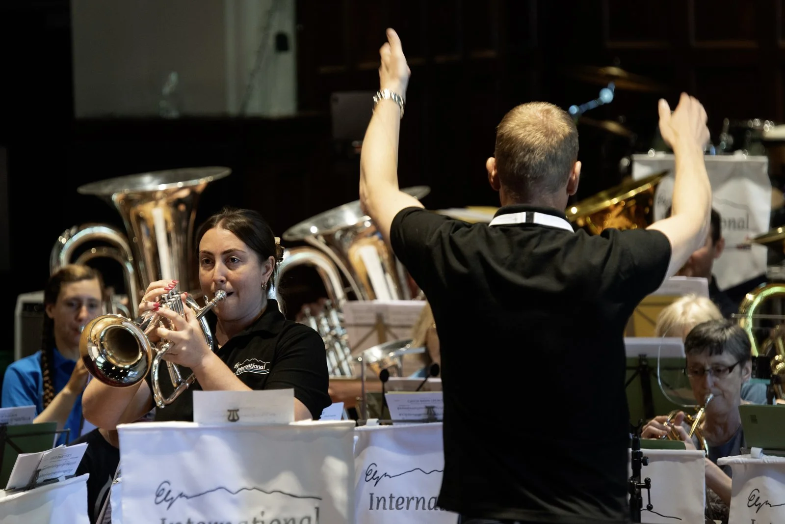 Orchestra performing with a conductor leading and members playing brass and woodwind instruments.