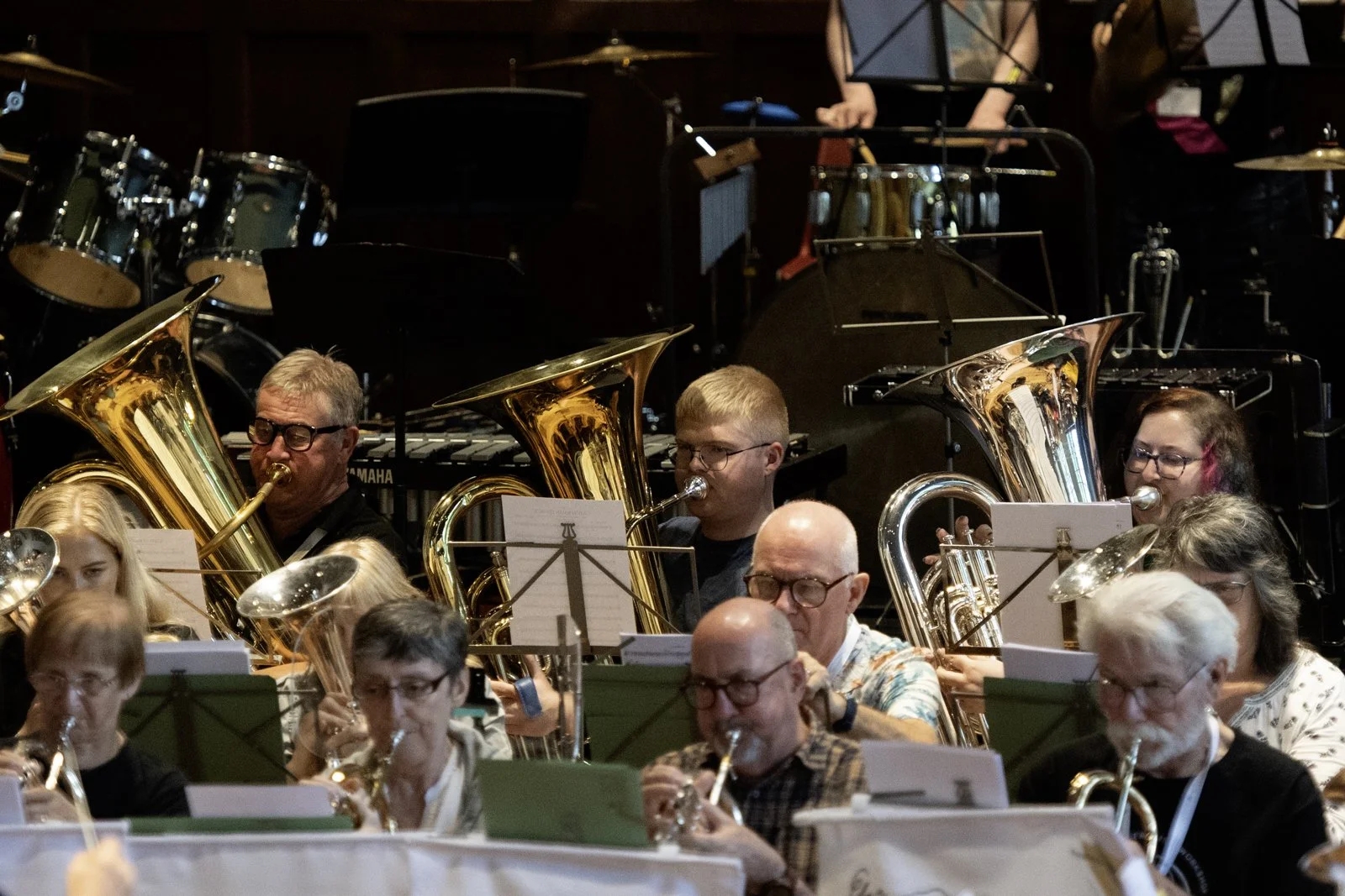 A large group of musicians playing brass and woodwind instruments during an orchestra performance. The musicians are seated with sheet music in front of them, and there are various percussion instruments and music stands in the background.