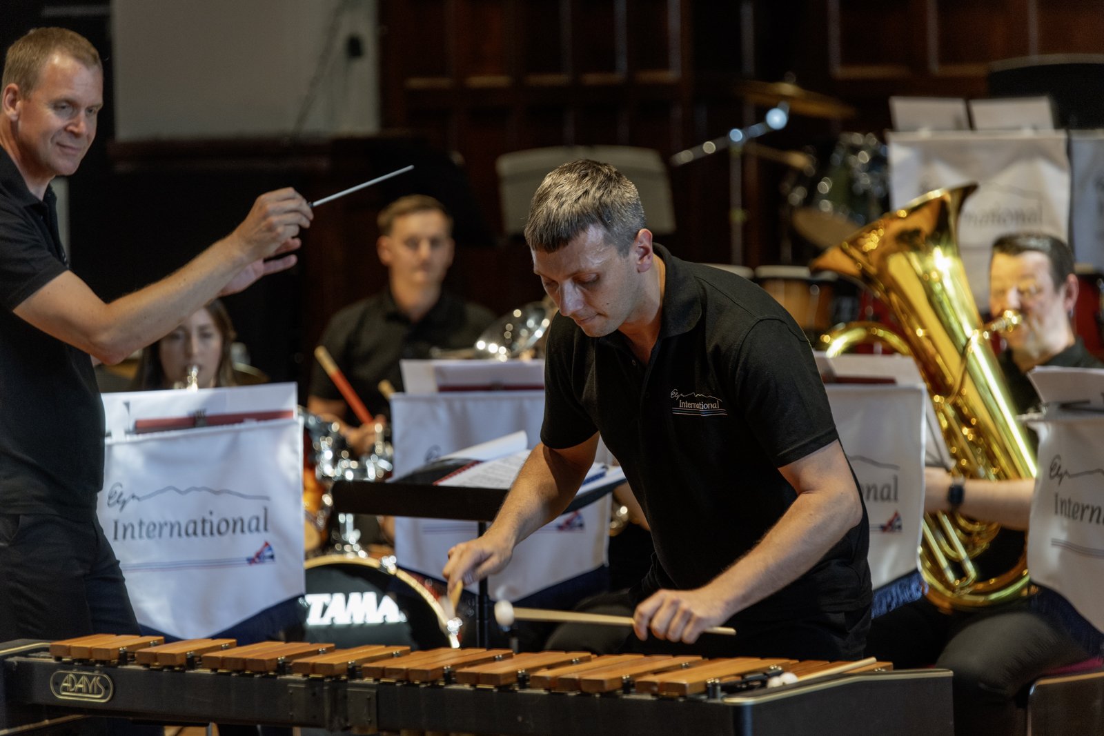 A man playing a marimba with a conductor leading an orchestra in the background, which includes musicians playing brass instruments.