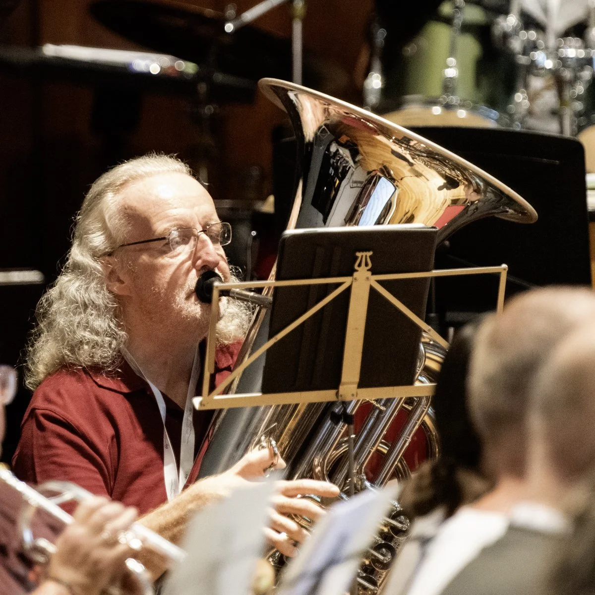 A man with long, curly gray hair and glasses is playing a tuba during an indoor musical performance.