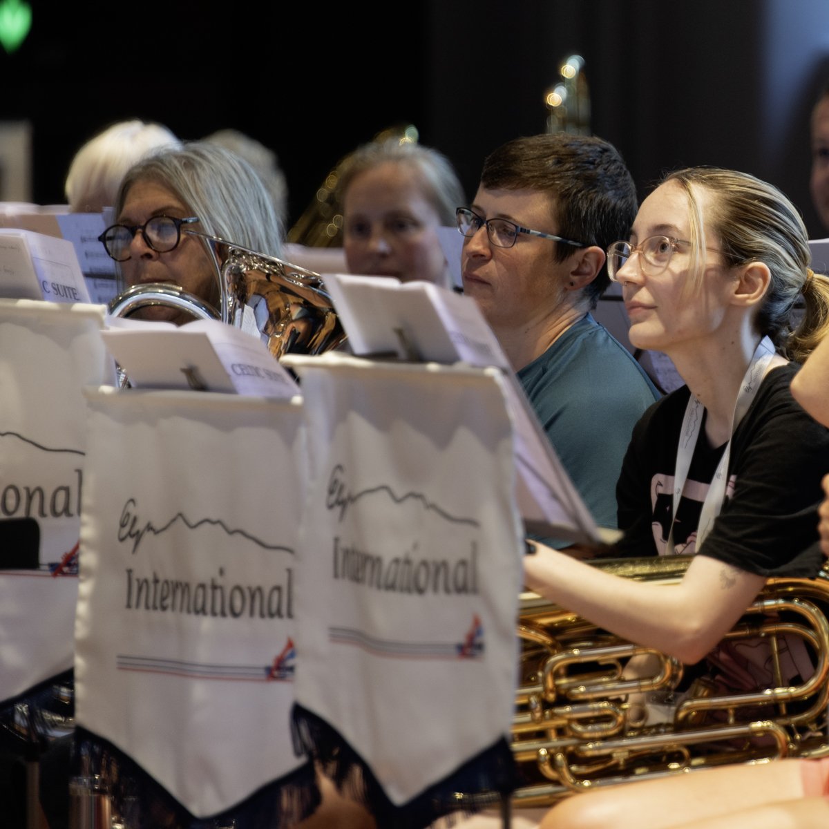 Members of a band sitting behind music stands labeled 'International' at a concert, with some playing brass instruments.